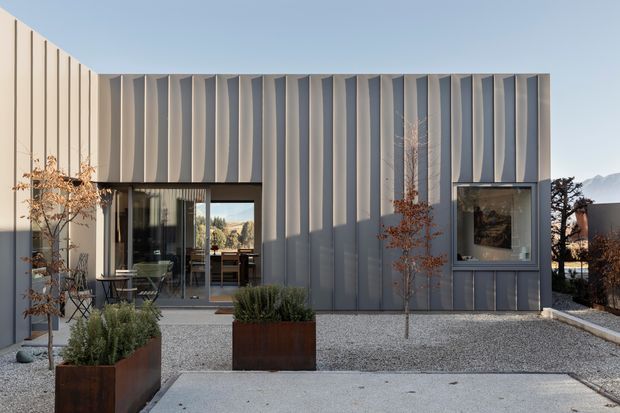 Modern single-storey building exterior with a sculptural vertically ribbed grey metal facade, large glazed openings, and a minimalist gravel courtyard. Rusted steel planter boxes with sparse landscaping sit in the foreground, while the clean geometric form and mountain landscape beyond reinforce the contemporary architectural character.