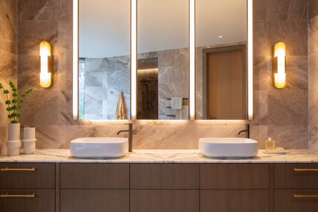 Luxury contemporary bathroom vanity with twin white vessel basins on a marble-look stone benchtop, dark timber cabinetry, illuminated full-height mirror panels, warm wall sconces, and a glimpse of a tiled shower area and timber door in the reflection.