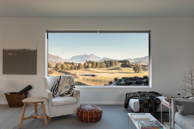 A bright, calm living room with a large picture window framing a rural mountain and valley landscape. The room has pale walls, light timber flooring, a neutral upholstered armchair and sofa, a patterned throw, a low round leather ottoman, a small timber stool, a woven basket, and a minimalist coffee table. The space combines warm natural textures with understated contemporary furnishings.