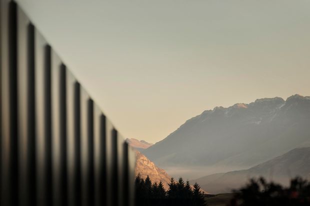 A minimalist architectural view showing a dark vertical-panel facade or screen receding diagonally from the foreground toward a dramatic mountain landscape. Warm, hazy light illuminates the distant peaks, while silhouetted trees and terrain sit along the lower edge. The composition contrasts the crisp, repetitive building element with the natural alpine setting.