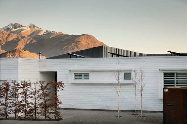 A modern single-storey residential facade with white horizontal cladding, narrow horizontal windows with exterior shutters, a dark timber entry door, and a dark low-pitched roofline. Sparse planted trees and gravel landscaping sit in front, with rugged sunlit mountains visible behind the home.