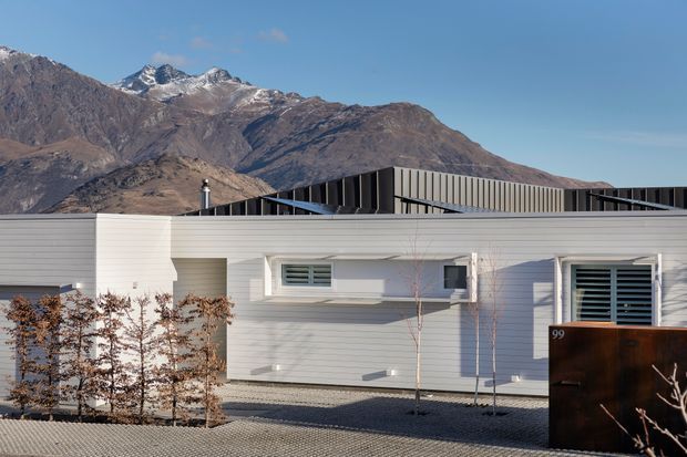 A low, contemporary single-storey building with pale horizontal cladding, crisp white window surrounds, dark louvred roof elements, and a sculptural dark entrance gate marked 99. The building sits in a minimalist gravel landscape, with rugged snow-dusted alpine mountains prominently visible behind it under a clear blue sky.