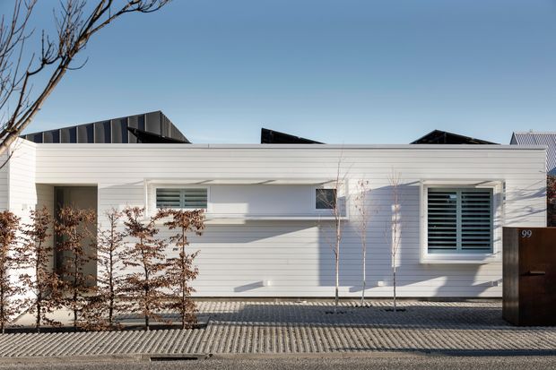 Street-facing view of a low, single-storey contemporary residence with a crisp white horizontal weatherboard facade, projecting flat and shallow pitched roof forms, deep-set openings, and prominent louvred windows. A paved forecourt and sparse planted screening sit in front of the house, with neighbouring rooflines visible behind.