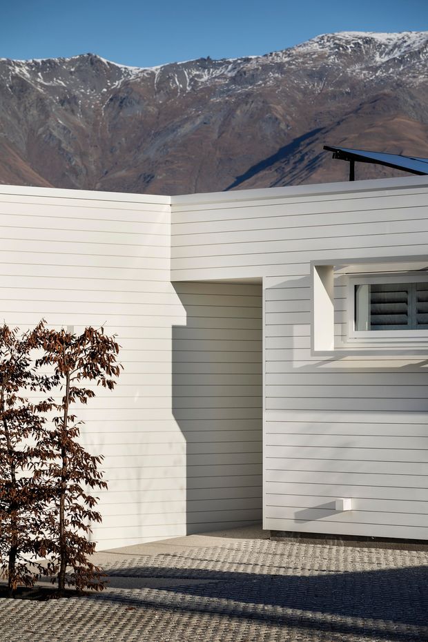 Exterior detail of a minimalist, low-profile house with white horizontal weatherboard cladding, a projecting box-like entry or courtyard wall, and a small recessed window with dark framing. Patterned paved ground and a young tree sit in the foreground, while a dramatic snow-dusted mountain range forms the background. Strong sunlight creates crisp architectural shadows.