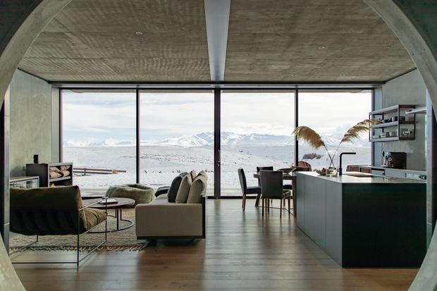 A contemporary open-plan living, dining and kitchen interior framed by exposed concrete arches and a textured concrete ceiling. Full-height glazing provides expansive views of a snow-covered mountainous landscape. The room features dark timber flooring, a sculptural dark kitchen island with open shelving, a dining table, and relaxed contemporary seating in muted earthy tones.