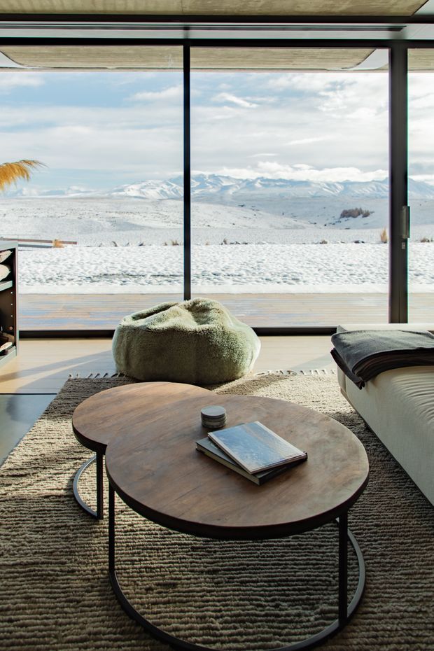 A warm, contemporary living room overlooking a snow-covered mountainous landscape. Floor-to-ceiling black-framed glazing opens the room visually to the alpine setting. The interior features a large textured neutral rug, an organic rounded timber coffee table with dark metal legs, a soft sculptural greenish lounge chair, and part of a light upholstered sofa. Natural light, timber decking, and muted earthy finishes create a relaxed, refined atmosphere.