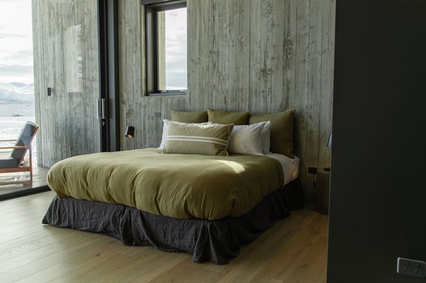 A moody, minimalist bedroom with a low-profile bed dressed in olive-green bedding, layered neutral pillows, and a dark charcoal bed skirt. The room features vertically textured concrete-look walls, warm timber flooring, dark window and door frames, and expansive glazing opening toward a mountainous landscape and outdoor seating. A small wall-mounted reading light sits beside the bed.