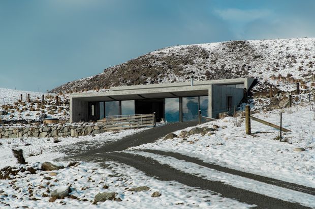 A low, contemporary concrete house is set into a snow-covered, rocky hillside. The building has a broad flat roof, dark window and door frames, extensive glazing, and a simple linear form. A paved driveway or walkway approaches the entrance, with stone retaining walls and rustic fencing around the exposed alpine landscape.