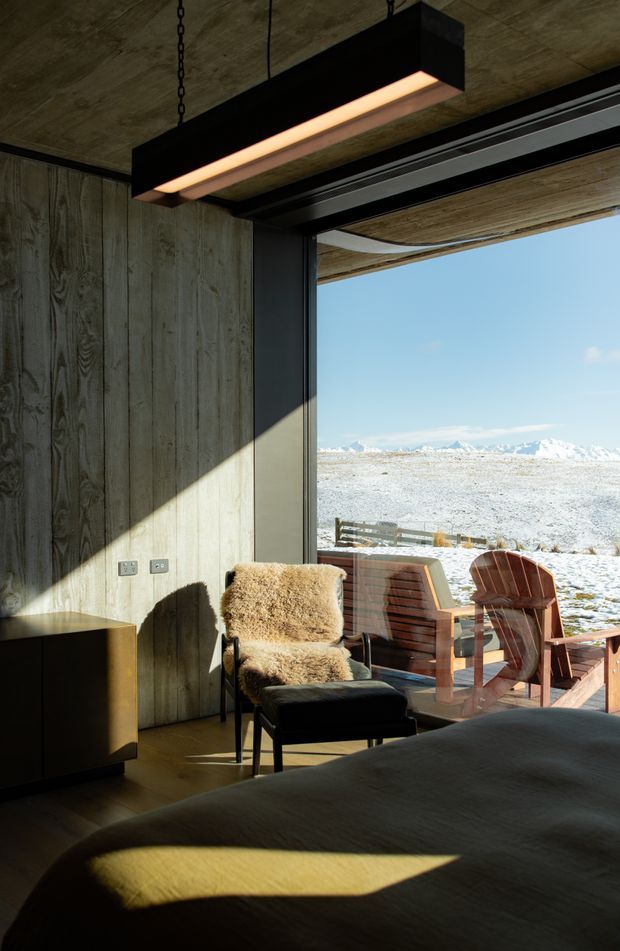A dark, refined bedroom with vertical timber wall panelling, a low-profile bed, leather and sheepskin lounge chair, black metal-framed cabinetry, and a suspended linear pendant light. Full-height sliding glazing opens to a timber deck with outdoor Adirondack-style chairs and a vast snow-covered alpine landscape beyond. Strong sunlight creates dramatic geometric shadows across the interior.