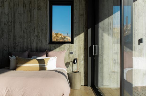 A compact, modern bedroom with a low-profile bed dressed in pale blush-pink bedding and layered neutral pillows. The room features dark textured concrete walls, warm timber flooring, a narrow black-framed window, and full-height black-framed glazed doors or windows with strong sunlight and reflections. A small black bedside lamp and cylindrical bedside table complete the restrained, contemporary interior.