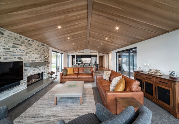 Spacious open-plan living room with a high pitched timber-lined ceiling, exposed central beam, grey carpet, and large area rug. The room features several brown leather sofas and armchairs, a pale upholstered coffee table, a dark stone fireplace with a natural rough-stone surround, built-in or freestanding timber media cabinetry, and extensive glazing. A kitchen is visible beyond the living area.