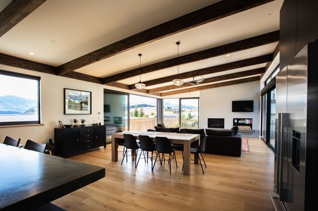 Open-plan contemporary living and dining area with a kitchen visible in the foreground. The room features exposed dark timber ceiling beams, light timber flooring, large windows and glazed doors with mountain views, a dining table with black chairs, and a living area with a fireplace and television.