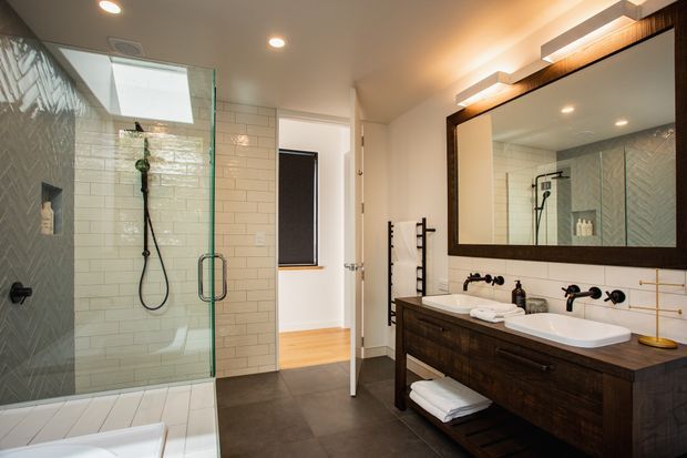 A contemporary bathroom with a large frameless glass shower enclosure, glossy white subway wall tiles, dark charcoal floor tiles, and a double-sink timber vanity. Matte black wall-mounted tapware and shower fittings contrast with the warm dark wood mirror frame and vanity. A freestanding towel ladder and recessed lighting complete the clean, refined space.