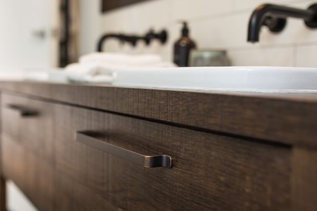 Close-up of a dark wood bathroom vanity with integrated drawers and slim metal handles, topped by a white basin. Matte black wall-mounted tapware and a folded white towel are visible against light tiled walls.