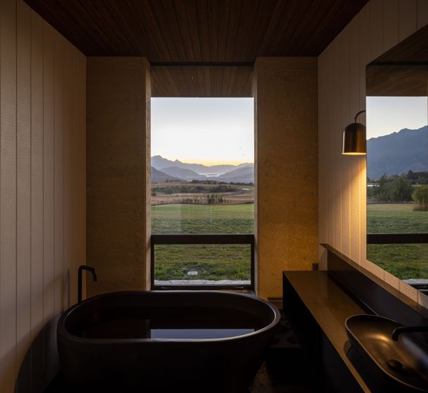 A dark, refined bathroom with a freestanding oval bathtub positioned in front of expansive windows overlooking a rural valley and mountains. The room features warm timber ceiling and wall panelling, textured earthy-toned vertical elements, a dark built-in vanity with a sculptural basin, and a suspended pendant light. The atmosphere is tranquil and luxurious, with strong indoor-outdoor visual connection.