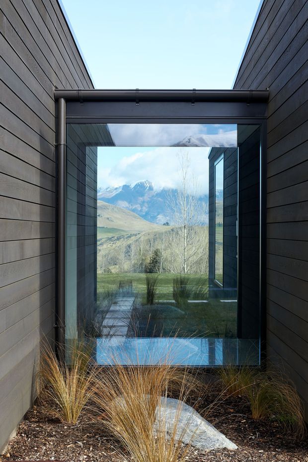 A narrow exterior courtyard or entry passage clad in dark horizontal timber boards, framed by tall side walls and a glazed overhead connection. Full-height glazing provides a strong view toward a rugged mountain landscape, with reflected sky and surrounding architecture. Ornamental grasses, dark mulch, and pale stepping stones form a minimalist landscaped foreground.
