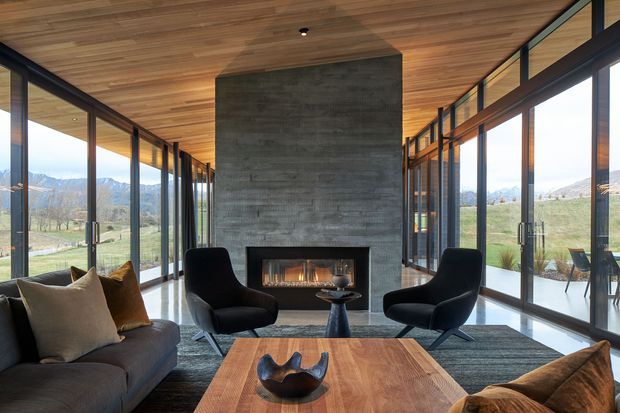 A contemporary living room with floor-to-ceiling glazing on both sides, a central textured concrete fireplace wall, timber-lined ceiling, dark lounge furniture, and a large solid timber coffee table. The space opens to expansive mountain and rural views.