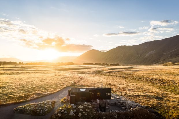 A small dark-clad building sits within a landscaped rural valley, surrounded by open fields, curving paths, planted garden beds and large mountain slopes. Warm low sunlight creates a dramatic golden-hour scene.