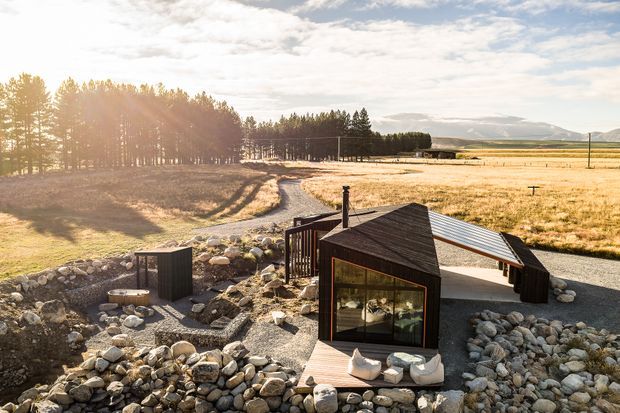 Aerial view of a compact, modern cabin-like building set within a rocky landscaped site beside open rural fields and a stand of trees. The structure has dark timber cladding, a steep asymmetrical roof, large glazed openings, and a lighter corrugated metal roof section. Gravel paths, stone landscaping, and a small outdoor seating area surround the building.