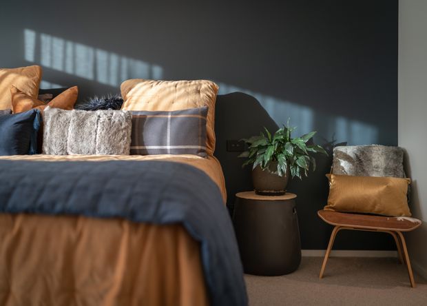 A warmly styled bedroom with a partially visible bed dressed in layered tan, navy, grey, and textured cushions, set against a charcoal feature wall. A small round dark bedside table holds a leafy plant, while a tan leather sling chair with a grey cushion sits nearby. Strong sunlight casts window-grid shadows across the wall, creating a cozy, refined atmosphere.