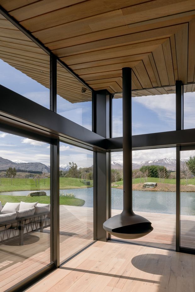 A contemporary glazed living space with expansive floor-to-ceiling windows overlooking a landscaped pond and snow-capped mountains. The room features a warm timber-lined ceiling and timber flooring, dark slender structural frames and columns, and a sculptural suspended black fireplace. A partially visible outdoor lounge sits beyond the glazing.