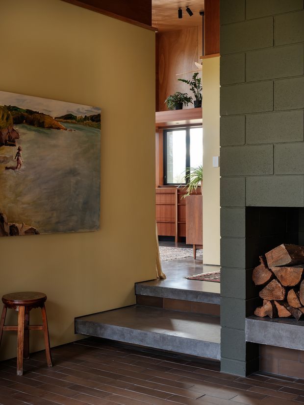 Warm, moody interior view looking through a stepped opening toward a sunlit living area. The foreground features dark brick-like floor tiles, a sculptural painted wall with artwork, a timber stool, and a substantial dark masonry fireplace with stacked firewood. Beyond are timber-lined walls and ceiling, built-in timber cabinetry, indoor plants, and large windows. The space has a curated mid-century-inspired character with strong natural timber and masonry detailing.