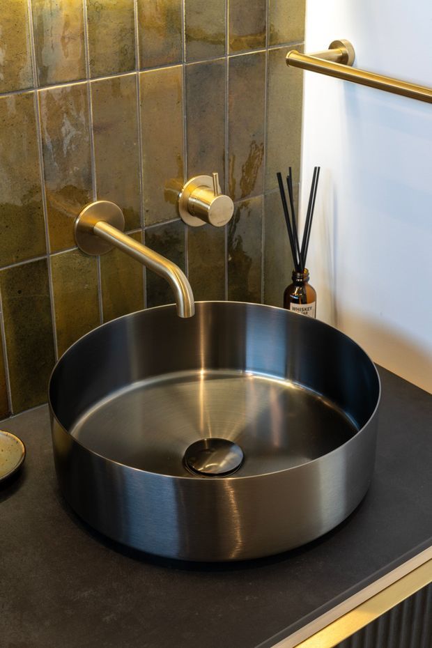 Close-up of a contemporary bathroom vanity featuring a large round brushed metal vessel basin on a dark countertop, wall-mounted brass/gold tapware, and a dark mottled rectangular tile splashback. A horizontal brass towel rail and diffuser sit against the white wall.