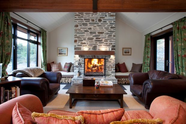 A warm, traditional living room centred around a large exposed-stone fireplace with a lit fire and timber mantel. Leather armchairs and sofas, patterned cushions, floral curtains, timber ceiling beams, and a dark wood coffee table create a cosy lodge-like atmosphere. Large windows and glazed doors provide views to greenery outside.