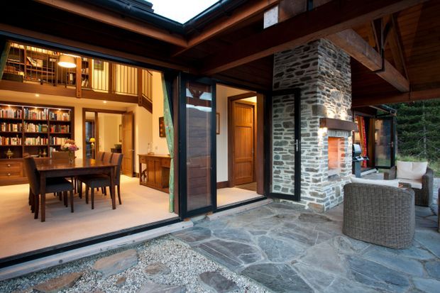 Covered courtyard or terrace opening into a warmly lit dining and living area. The design features exposed timber beams and ceiling structure, dark timber-framed glazing and doors, a substantial rough-stacked stone fireplace or chimney wall, slate or irregular natural-stone paving, and built-in timber shelving filled with books. Wicker outdoor lounge furniture sits beside the fireplace, creating a rustic, sheltered indoor-outdoor space.