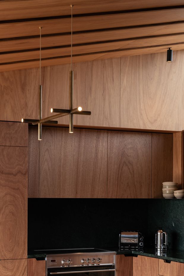 A refined, warm-toned kitchen interior featuring extensive dark timber veneer cabinetry, a black stone benchtop and splashback, and a timber-lined ceiling with dark recessed channels. A stainless-steel freestanding range, toaster oven and kettle sit beneath the cabinetry. Slim contemporary pendant lights and a small black ceiling spotlight provide focused illumination. Ceramic bowls are displayed on an open shelf at the right.
