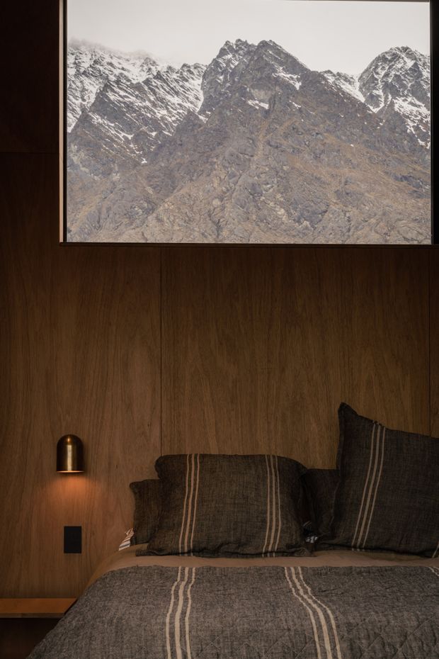 A moody, intimate bedroom with dark timber wall panelling and an integrated bedside shelf. A warm, compact wall sconce illuminates the bed, which is dressed in charcoal-grey striped bedding and cushions. Above the bed, a large framed mountain photograph or view creates a strong connection to the alpine landscape.