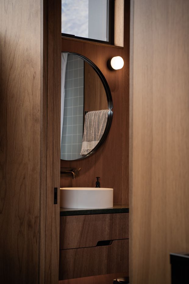 A compact, warmly lit bathroom vanity viewed through a partially open timber door. The space features dark wood wall panelling and built-in vanity cabinetry, a black stone benchtop, a white vessel basin, black tapware, a large rounded black-framed mirror, and a small spherical wall light. A tiled shower or wet-area surface is visible in the mirror reflection.