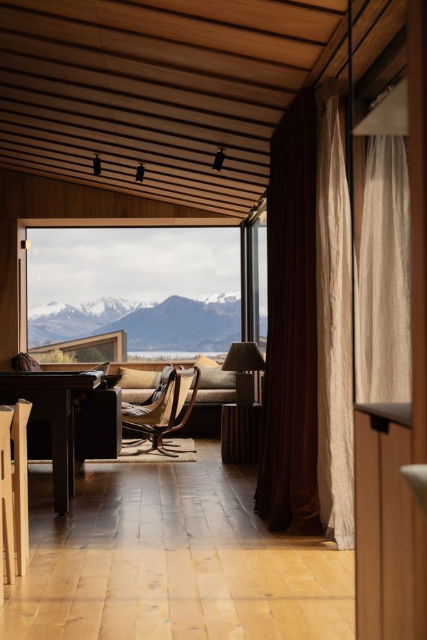 Warm, wood-lined interior looking through a living room toward expansive glazing and a dramatic mountainous landscape. The room features a pitched slatted timber ceiling with black track lighting, timber wall and floor finishes, dark curtains, a low sofa, mid-century-style lounge chairs, and a dark pool or billiards table in the foreground. The setting feels like a refined mountain retreat with strong indoor-outdoor connection.