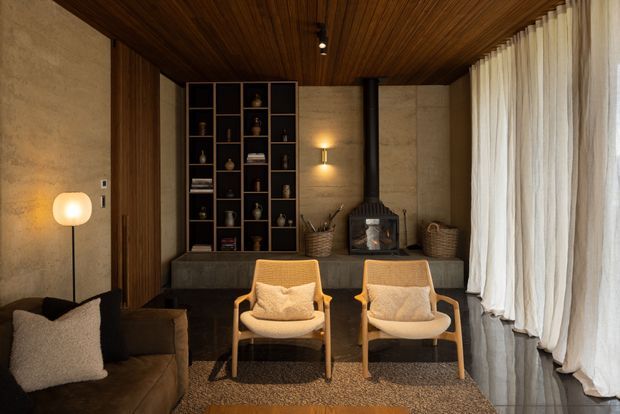 Warm, refined living room with a dark timber slatted ceiling, textured concrete-toned walls, dark built-in shelving, a black wood-burning stove, lounge seating, and full-height sheer curtains. The room combines natural materials, soft lighting, and mid-century-inspired armchairs in a calm, cozy setting.