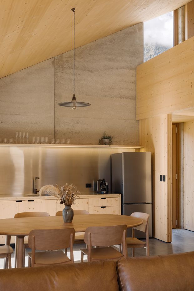 Warm contemporary open-plan kitchen and dining area with extensive pale timber lining, a textured concrete feature wall, pale timber cabinetry, stainless-steel refrigerator, and a large oval timber dining table. A leather sofa occupies the foreground, while a high window and timber-framed opening reveal a glimpse of the surrounding landscape.