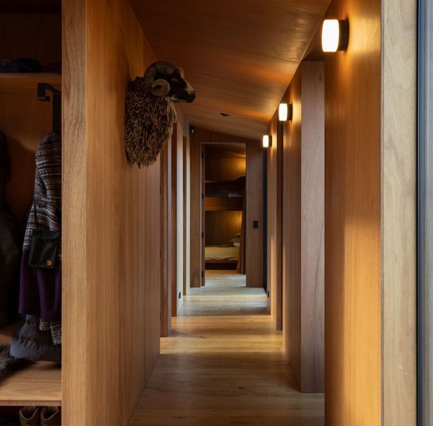 A narrow, warmly lit timber-lined hallway extending through the home, with continuous timber flooring, walls and ceiling, recessed doorways, wall-mounted cylindrical sconces, and built-in wardrobe storage visible at the left. A mounted ram head adds a rustic decorative detail, while a bedroom or sleeping area is visible at the far end.