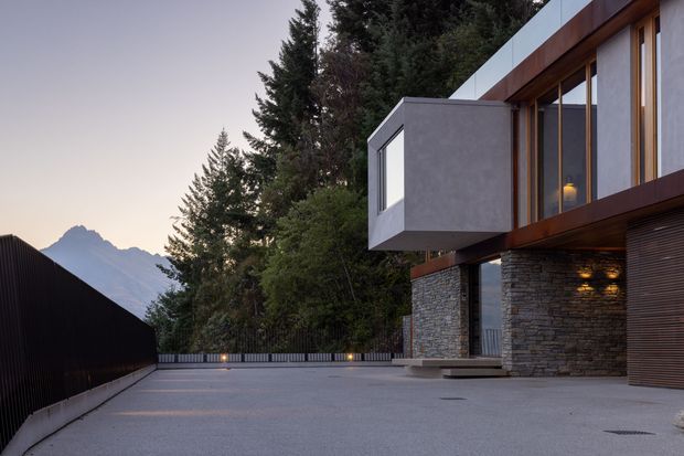 Exterior architectural photograph of a contemporary multi-level residence set against a wooded mountain landscape. The building features dark stone masonry, extensive vertical glazing, warm timber-toned cladding, a projecting box-like upper volume, and a broad paved forecourt or driveway. The image is taken outdoors and focuses on the facade and entry-side exterior.