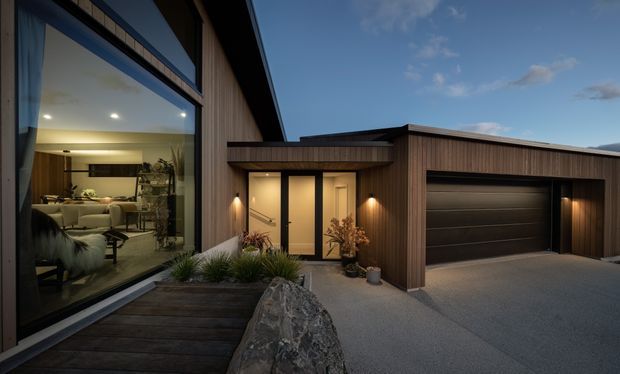 Contemporary single-storey home exterior at dusk, featuring dark vertical timber cladding, a dramatic floor-to-ceiling glazed living area, a recessed entry, and an integrated double garage. A timber deck, stone boulder, and low ornamental planting form the foreground landscaping.