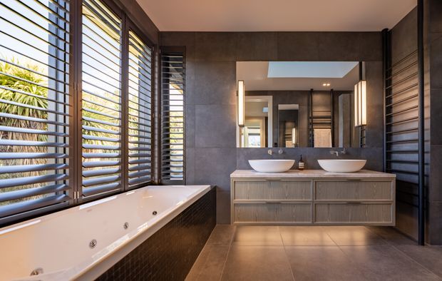 Contemporary bathroom with a freestanding-style white spa bath beside large timber louvred windows, a double floating timber vanity with vessel basins, and a wide mirrored wall. Dark large-format tiled walls and flooring create a refined, warm-toned interior with strong natural light and views of tropical planting through the shutters.