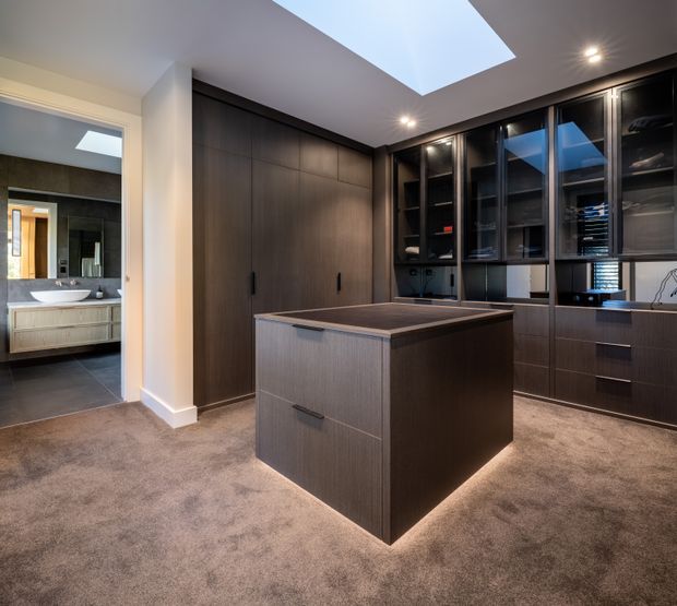 A sophisticated walk-in wardrobe with dark timber built-in cabinetry, glass-fronted shelving, drawers, and a central island. The room has plush grey carpet, integrated lighting, and a large skylight. A bathroom with a vanity and basin is visible through the open doorway at left.