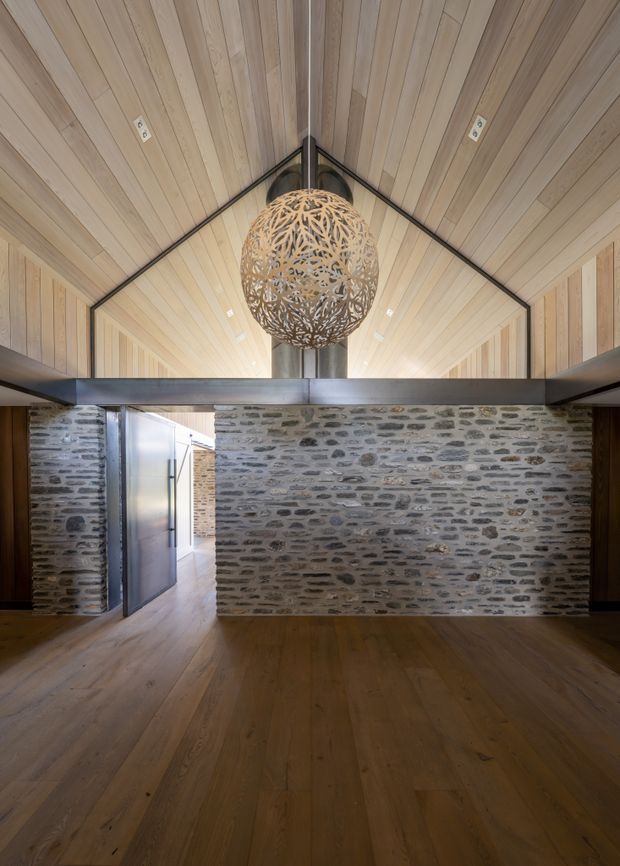 Interior view of a dramatic gabled space with a high timber-lined pitched ceiling, exposed dark structural beams, and a large spherical woven pendant light. A substantial rough stone masonry wall divides the room, with a doorway opening to a corridor on the left. The floor and ceiling use warm timber boards, creating a rustic yet refined architectural atmosphere.
