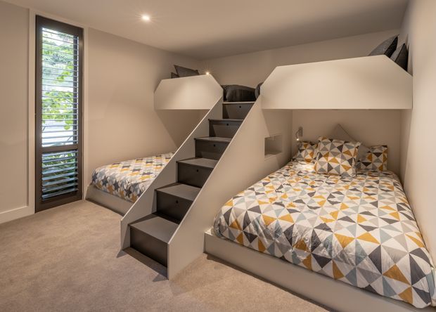 Compact shared bedroom with a clever built-in bunk-bed arrangement. Two lower beds sit beneath enclosed upper bunks, connected by a central staircase with dark stair treads and integrated storage. Neutral painted walls, beige carpet, recessed ceiling lighting, and a tall louvred window create a warm, functional interior. Geometric bedding adds muted mustard, grey, white, and taupe accents.