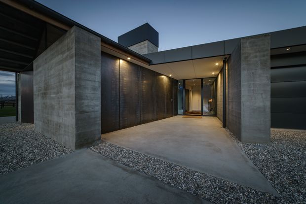 Dusk exterior view of a contemporary residence entry and covered driveway approach. The composition features large raw concrete blade walls, dark metal cladding and garage doors, a flat roof, recessed lighting, a concrete forecourt, and gravel landscaping. A glazed entry is visible at the end of the passage.
