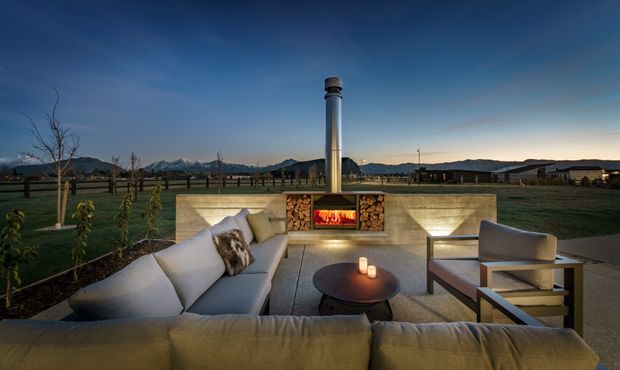 An outdoor sunken lounge courtyard at dusk, arranged around a built-in stone fireplace with a tall metal chimney. The space includes a large upholstered sectional sofa, two lounge chairs, a round coffee table with candles, concrete or stone retaining walls, and open views toward a rural landscape and mountains.