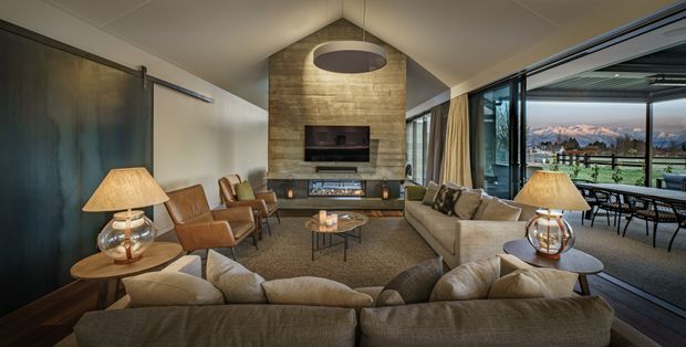 A sophisticated living room arranged around a stone-clad fireplace and television, with a large sectional sofa, leather lounge chairs, coffee table, and table lamps. Full-height sliding glass doors open to a covered outdoor terrace with dining furniture and expansive mountain views. The room has a warm, refined atmosphere with dark wall panels, neutral upholstery, and a large circular pendant light.