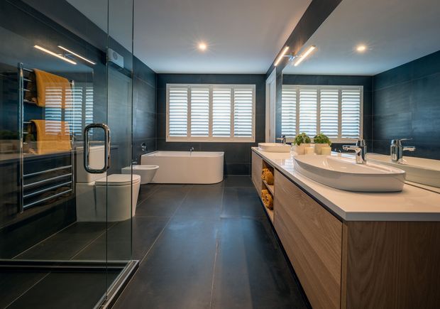 A spacious contemporary bathroom with dark tiled walls and floor, a freestanding white bathtub, glass-enclosed shower, double vanity with timber cabinetry and white vessel basins, wall-mounted mirrors, heated towel rail, and plantation shutters. The room is well lit with recessed and linear feature lighting.
