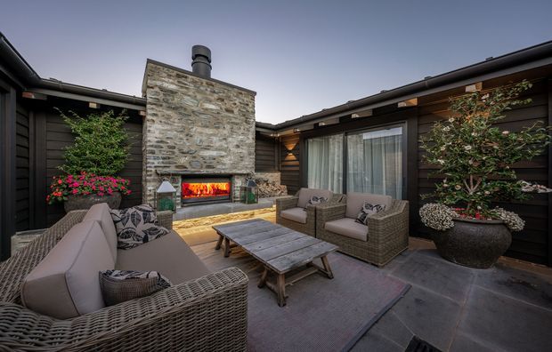 A sheltered outdoor courtyard lounge with a built-in stone fireplace, firewood storage, dark horizontal wall cladding, timber-lined eaves, woven outdoor seating, a large coffee table, potted plants, and glazed doors opening to the interior. The space has a warm, intimate resort-like atmosphere and is photographed at dusk.