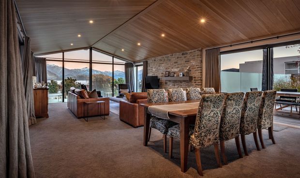 A spacious open-plan living and dining room with a warm timber-lined sloping ceiling, exposed stone feature wall, carpeted floor, large corner glazing, and mountain views. The room includes a substantial timber dining table with patterned upholstered chairs, leather lounge chairs, a fireplace or media wall, and heavy curtains. The interior has a refined lodge-like character with strong connection to the landscape.