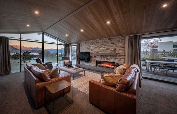 Spacious contemporary living room with a warm timber-lined sloping ceiling, expansive floor-to-ceiling glazing, brown leather sofas and armchairs, a textured natural-stone feature wall, and a built-in fireplace with visible flames. The room opens to a deck and overlooks a mountainous landscape.