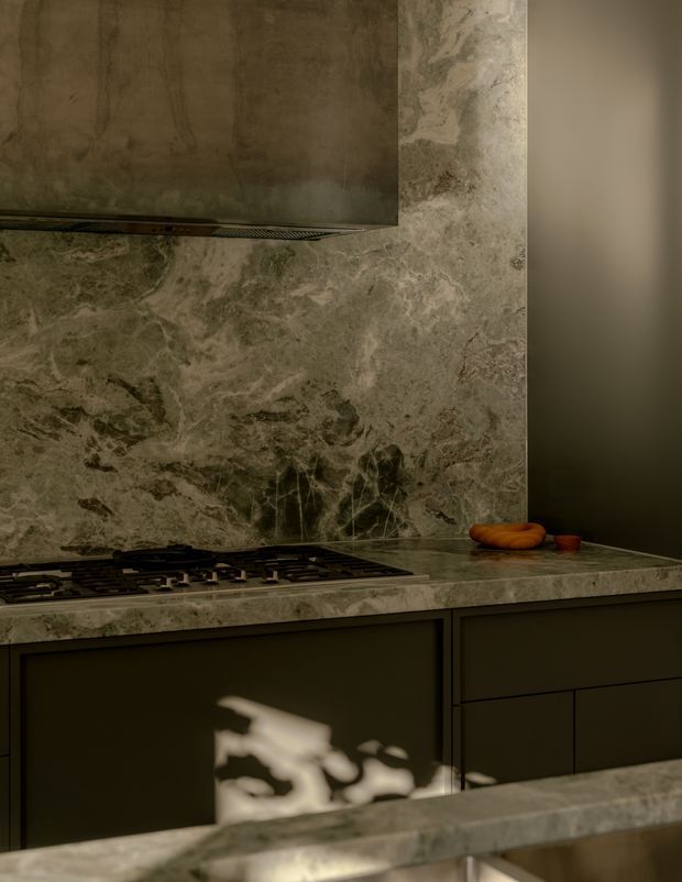 Moody close-up of a contemporary kitchen featuring dramatic veined grey-green natural stone across the splashback and benchtop, dark matte cabinetry, and a built-in gas cooktop beneath a matching stone or metal rangehood. Warm sunlight creates strong shadow patterns across the cabinetry and foreground island.